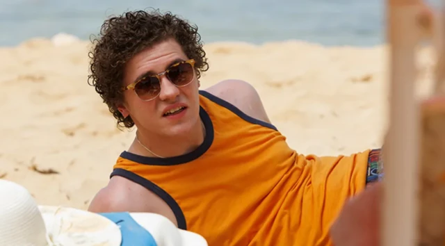Un joven con camiseta de tirantes naranja y gafas de sol parece hacer ademán de incorporarse de la playa en la que está tendido. 