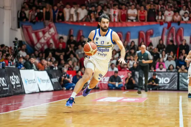 Sebastián Vega toma la pelota de básquetbol con su mano derecha en medio de una jugada; viste la indumentaria alternativa, que es preponderantemente blanca con franjas azules y amarillas.