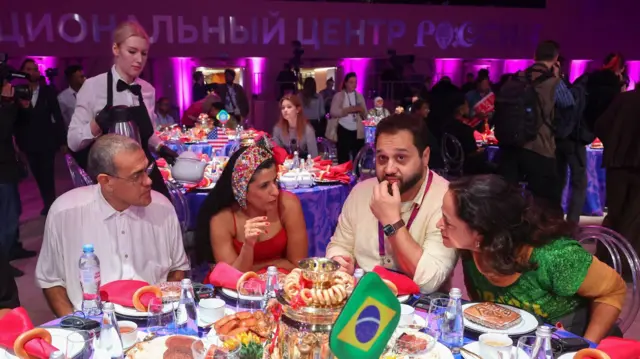 En lo que parece ser una gran reunión, vemos a cuatro personas sentadas en una mesa que está repleta de una variedad de alimentos y bebidas. Entre los platos hay una pequeña bandera brasileña. En el fondo, se ven meseros, fotógrafos, más personas sentadas y conversando y un leterero en alfabeto cirílico.