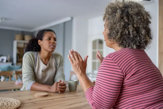 Dos mujeres sentadas a una mesa. Una mujer levanta sus manos para detener la conversación de la otra