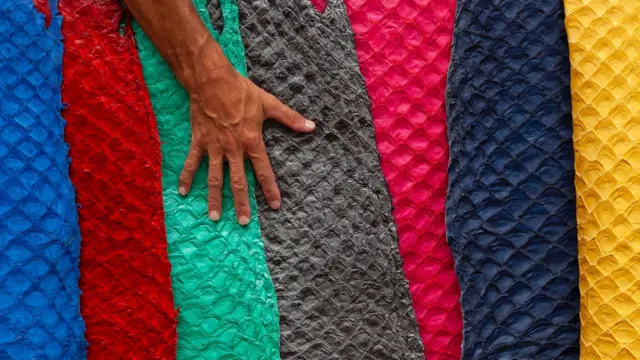 Una mano extendida sobre rollos de piel de pirarucu teñida de colores vivos: azul, rojo, verde, gris, rosa, azul oscuro y amarillo.