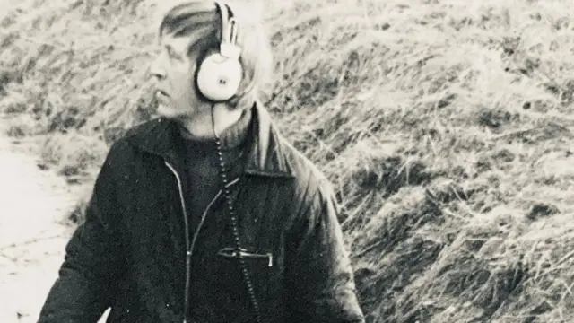 Una imagen en blanco y negro de Dave Derby, que data de la década de 1970 aproximadamente. Mira hacia la izquierda de la imagen y lleva puestos unos auriculares con cable y una chaqueta medio abrochada sobre un jersey de cuello alto. Detrás de él se ve el borde cubierto de hierba de una zanja.