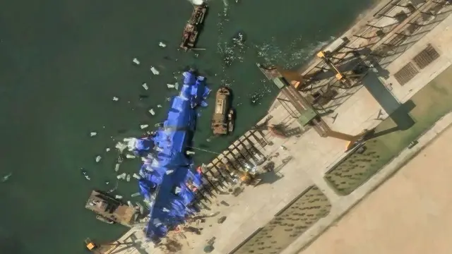 Imagen aérea por satélite de un barco en el puerto cubierto de tela azul, rodeado de embarcaciones más pequeñas.