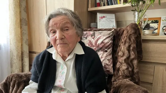 Wanda Traczyk-Stawska, de 98 años, mira directamente a la cámara. Está sentada en un sillón con una blusa bordada y un cárdigan.