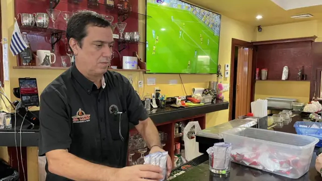Fernando Scariato detrás del mostrador en la parrillada El Leño. Viste una camisa negra, y detrás de él se puede ver una pequeña bandera uruguaya. A la derecha de la foto, de fondo, se ve un televisor transmitiendo un partido de fútbol.