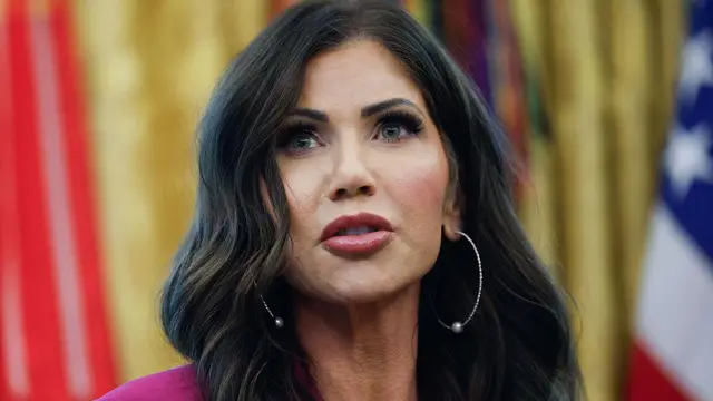 Primer plano de Kristi Noem, secretaria de Seguridad Nacional, con unas banderas difuminadas detrás de ella.