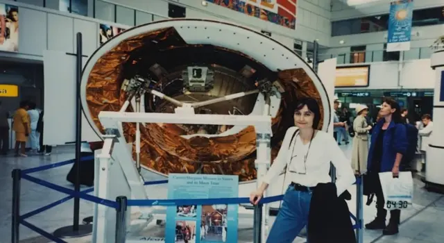 Michele Dougherty, de 30 años, con un top blanco y vaqueros azules, gafas colgando del cuello y una chaqueta negra colgada del brazo. Está de pie delante de una parte redonda de una nave espacial, en parte blanca pero con partes de lámina de cobre. Está delimitada por una barandilla azul. La escena parece transcurrir en un museo, o tal vez en un aeropuerto, y hay gente alrededor.