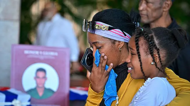 Una mujer se seca las lágrimas mientras abraza a una niña, frente a la fotografía de un soldado 