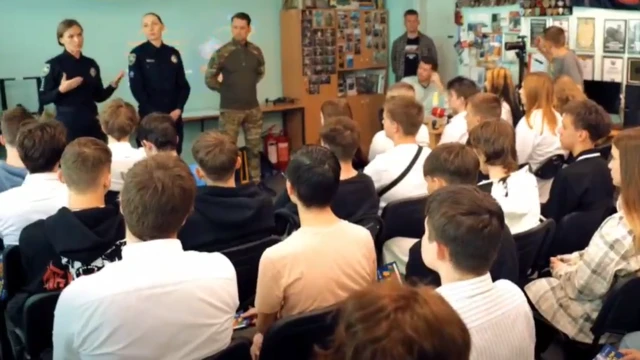 Una agente del SBU, vestida con uniforme, se dirige a una clase de adolescentes en una escuela de Kyiv sobre los peligros del reclutamiento en línea. Junto a ella se encuentran otros dos agentes del SBU, un hombre y una mujer, y un profesor. La charla está siendo grabada por un camarógrafo desde un lado del aula.