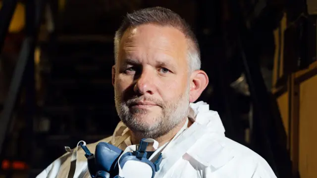Fotografía de cerca de Ben Giles. Tiene 49 años y cabello corto y canoso. Lleva una mascarilla alrededor del cuello. Se puede ver su EPI, un traje blanco con capucha, alrededor del cuello.