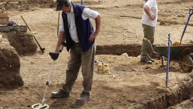 Dave Derby con su detector de metales en la granja Whitehall durante la excavación de 2003. Está inclinado mirando al suelo y sosteniendo el detector con la mano derecha. Detrás de él se ven zanjas y terreno despejado.