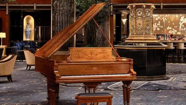 Un piano de cola de madera marrón en el centro, detrás un antiguo reloj dorado y butacas de madera con tapizado color crema.
