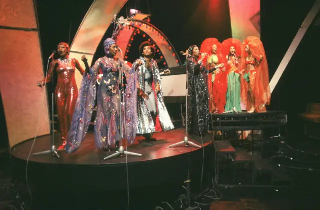 Una vibrante presentación musical con un grupo diverso de artistas vestidos con deslumbrantes trajes con lentejuelas, capas y tocados ornamentados.