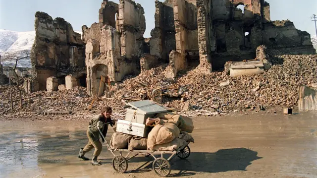 Un joven afgano empuja una carreta cargada de sacos y cajas por una calle embarrada. Detrás de él se ve un edificio destruido y escombros.