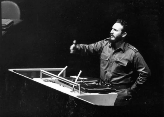 Imagen en blanco y negro de Fidel Castro hablando de pie desde el podio de oradores de la Asamblea General. Vestido con lo que parece un uniforme militar, Castro tiene su mano izquierda apoyada sobre la cintura y la derecha levantada en el aire mientras habla.