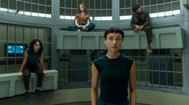 Una mujer joven en pie. Detrás de ella, otros tres jóvenes están sentados en bancos. Dos de ellos, en un banco elevado, parecen dormitar