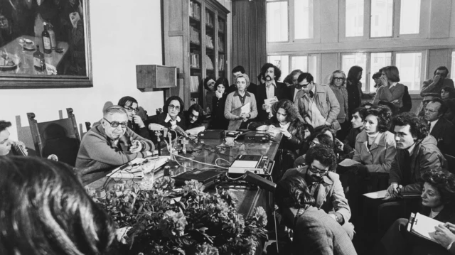 Sartre sentado en un escritorio con micrófonos al frente y varios periodistas mirándolo en un salón