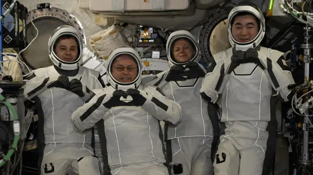 Cuatro astronautas, vestidos con trajes espaciales blancos y cascos, posan para las cámaras sonriendo y haciendo gestos de corazón con las manos.