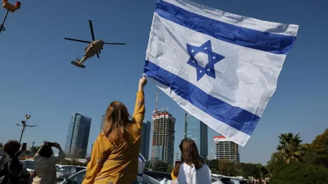 Helicópteros militares con rehenes a bordo descienden hacia el hospital. Una mujer en tierra los saluda agitando una bandera de Israel. 