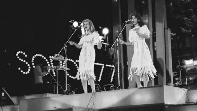 Dos mujeres con vestidos claros cantan en el escenario con micrófonos. En el fondo, bombillos de luz forman de logotipo de la edición de 1977 del festival Intervisión, llevada a cabo en la ciudad polaca de Sopot. La imagen es en blanco y negro.