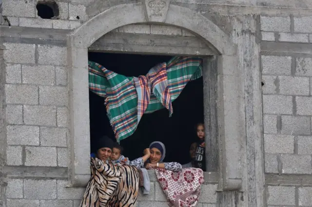 Mujeres y niños miran por la ventana de un edificio de piedra. Hay alfombras que cubren la ventana y cuelgan del techo.