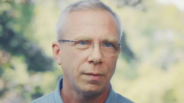 Benoit lleva gafas sin montura, tiene los ojos azules, el pelo gris y viste una camisa azul. 