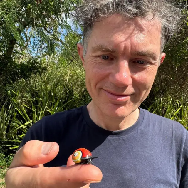 Un hombre sonriente con una camiseta azul marino sostiene un caracol de colores brillantes en un dedo