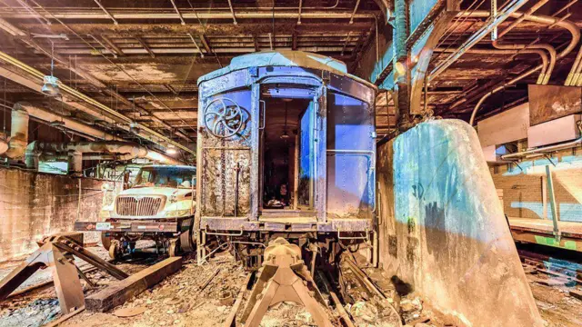 Un viejo vagón de tren que luce oxidado sobre unas vigas, en un túnel sucio lleno de escombros en el suelo. La parte delantera de un camión se ve en el fondo.