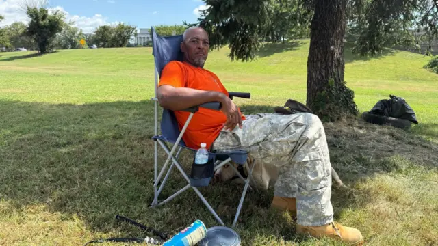 Un hombre con una camiseta roja y pantalones de camuflaje está sentado en una silla de camping azul a la sombra de un árbol, sobre un trozo de césped. Se puede ver a su perro durmiendo a su lado, debajo de la silla.