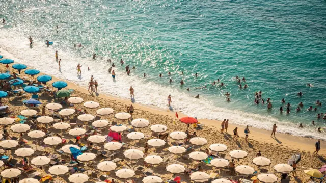 Una vista aérea de una playa en italia. Se ven decenas de sombrillas de playa y decenas de personas tanto en la arena como en el mar. 