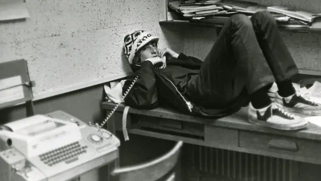 Un Bill Gates adolescente con sombrero, recostado sobre un escritorio, apoyado en la pared mientras sostiene un teléfono fijo, en una fotografía en blanco y negro.