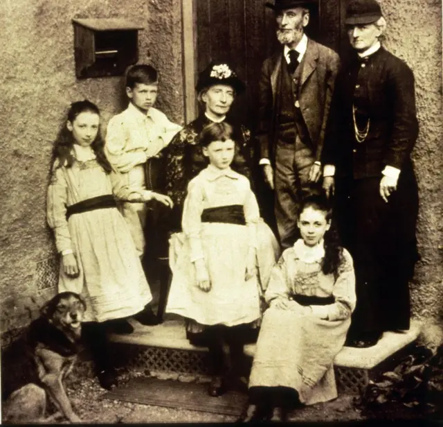 Eglantyne sentada en el escalón de la entrada de la casa con su tía y familiares