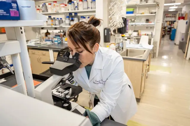 Tamar Gefen con una bata de laboratorio en el laboratorio observa una muestra en un microscopio 