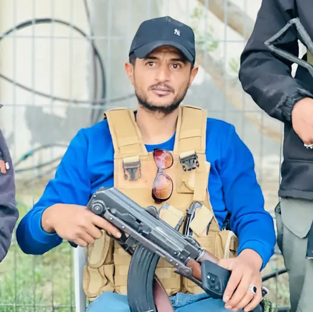 Yasser Abu Shabab viste una camisa azul, una gorra negra, un chaleco beige. Porta un fusil.