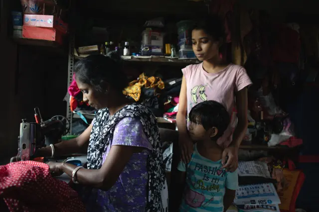 Mujer cosiendo tela roja en una máquina de coser dentro de una habitación pequeña y oscura, con estantes llenos de frascos y ropa en el fondo, mientras un niño y una niña permanecen de pie cerca.