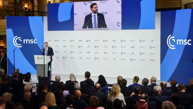 JD Vance, en un traje oscuro, se dirige a un público desde un podio, flanqueado por dos letreros azules que leen: 