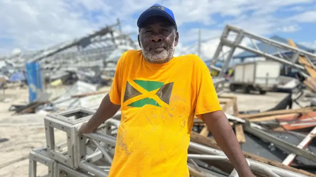 Jimmy Esson, entre los escombros de una casa, vistiendo una camisa amarilla con la bandera de Jamaica