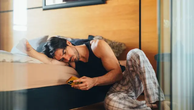 Hombre mirando el celular acongojado. Usa un pijama gris a rayas y una sudadera negra. 