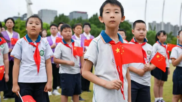 Un estudiante de primaria sostiene una bandera china en la mano. Detrás de él hay un montón de escolares con camisetas blancas y pantalones cortos azules con pañuelos rojos y azules y blancos en el cuello. Están de pie en un campo.