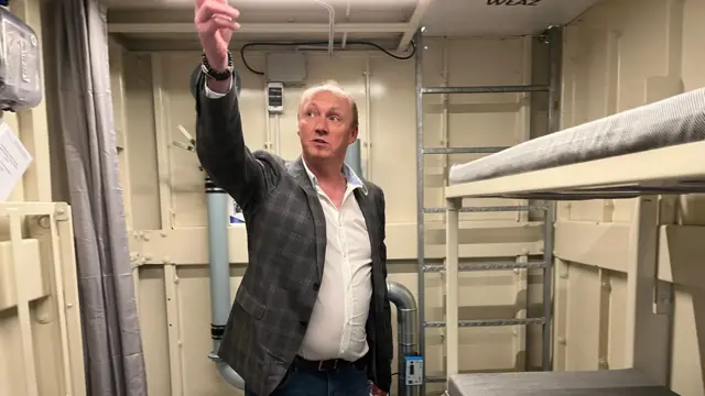 Janusz lleva una chaqueta a cuadros y vaqueros dentro de uno de sus refugios antiaéreos. Señala al techo. El refugio tiene una litera y un baño con cortina. Está limpio, pero escaso.