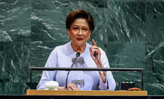 La primera ministra de Trinidad y Tobago, Kamla Persad-Bissessar, está parada en un podio y levanta una mano mientras habla.