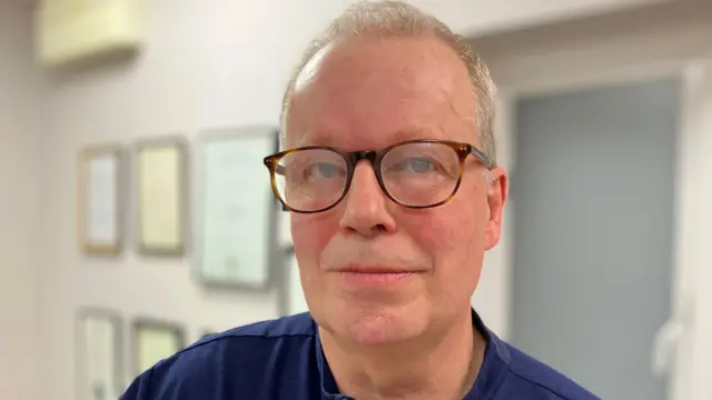 El doctor Paul Charlson tiene el pelo gris, gafas con montura de carey y lleva una camisa azul marino. Aparece en una foto dentro de una habitación pintada de blanco, con marcos colgados en la pared detrás de él.