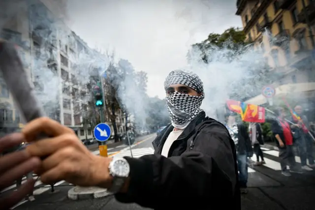Un hombre con la cara cubierta por un pañuelo palestino sujeta un palo y mira a la cámara en medio de las protestas en una calle de Turín, Italia. Se ve humo al fondo