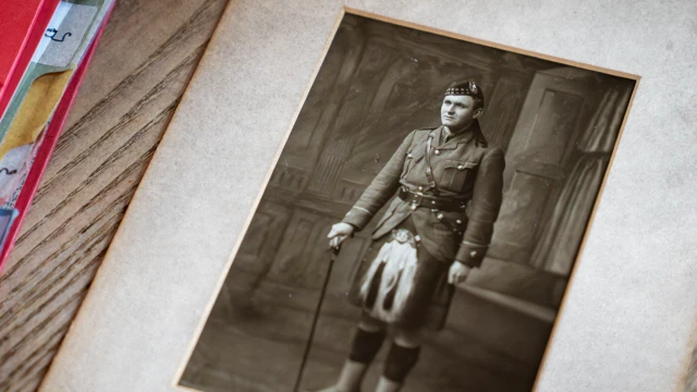 Fotografía en papel de James Grant Allan, de pie con falda escocesa, sporran y gorra militar. La fotografía es en blanco y negro, está enmarcada y se encuentra sobre una mesa de madera, con una carpeta roja a su izquierda. 