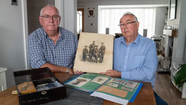 Imagen panorámica de dos hombres de unos 60 años sentados a una mesa de madera en una sala de estar, mientras sostienen una vieja foto sepia de cuatro hombres. Alistair, sentado a la izquierda, lleva una camisa azul a cuadros y Andrew, a la derecha, lleva una camisa azul claro. 