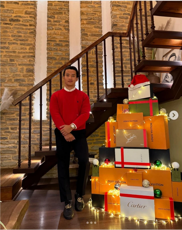 Saugat Thapa, con un suéter rojo y pantalones negros, se encuentra junto a un árbol de Navidad hecho con cajas de marcas de lujo.