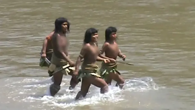 Tres mujeres de la tribu mashco piro caminan por las aguas poco profundas de un río. Llevan faldas hechas de materiales naturales, como hojas o fibras vegetales, y sostienen largas lanzas de madera. 