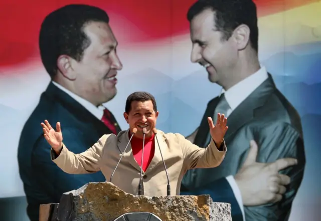 Hugo Chávez, delante de un estrado de piedra, con tres micrófonos. Tiene los brazos abiertos y sonríe. Va vestido con una camiseta roja y encima una chaqueta de traje de tela ligera. Al fondo, hay una gigantografía con una foto donde se ven a Chávez y Bashar al-Assad mirándose y sosteniéndose los brazosde modo amistoso y, como fondo de esa foto, los colores de las banderas de Siria y Venezuela. 
