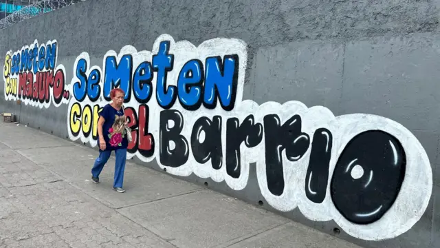 En una pared exterior gris de doble altura con alambre de púas en lo más alto, se extiende un letrero que dice 