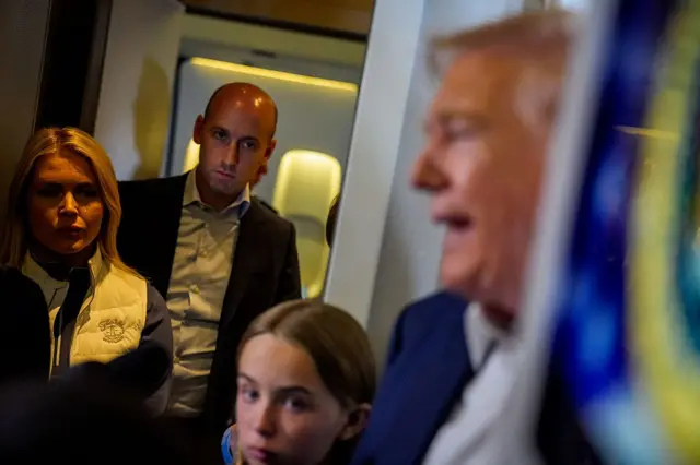 Trump, de perfil, habla con los periodistas en su avión presidencial, mientras una niña, su portavoz Karoline Leavitt y su asesor Stephen Miller lo escuchan desde un lateral.  